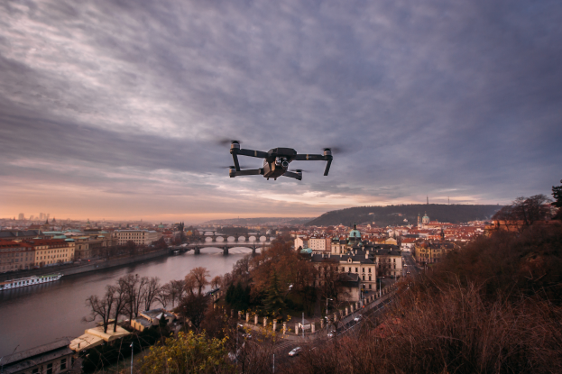 Ein Drohne fliegt über eine Stadt mit Gebäuden, Bäumen und Fahrzeugen, mit einem Fluss und einer Brücke im Hintergrund.