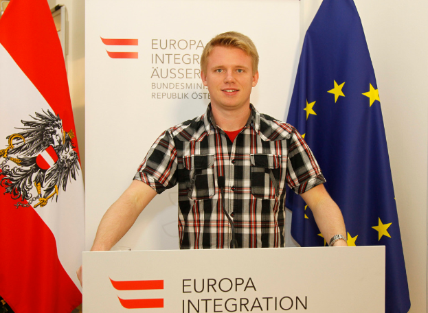 Ein Junge steht hinter einem Rednerpult mit einem Mikrofon, mit einem Plakat und Fahnen dahinter.