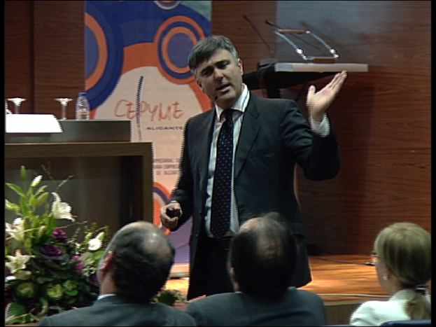 Ein Mann in einem schwarzen Blazer steht an einem Podium mit Mikrofonen, spricht in ein Mikrofon, mit einem Blumentopf oder einem Blumenstrauß in der Nähe, während drei Personen in Stühlen vor ihm sitzen, ein Tisch mit einem Buch, einer Wasserflasche und anderen Gegenständen daneben, vor einer braunen Wand und einer bunten Tafel mit Text im Hintergrund, wahrscheinlich in einem Konferenzsaal.