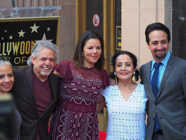 Eine Gruppe von Menschen auf einem roten Teppich, die lächeln und für ein Foto posieren, mit einer Tafel, einem Glaspodest und einer Wand im Hintergrund und einer Kamera auf der linken Seite.