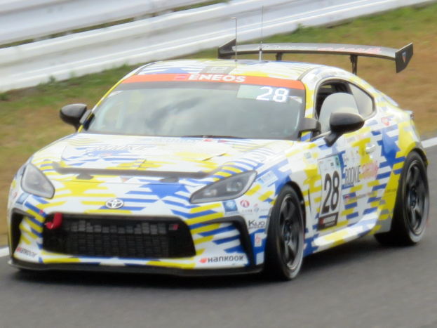 Ein weißer Toyota Supra GT3-Rennwagen mit Sponsorentext und Nummern auf einer Rennstrecke, von einem Geländer und Gras umgeben.