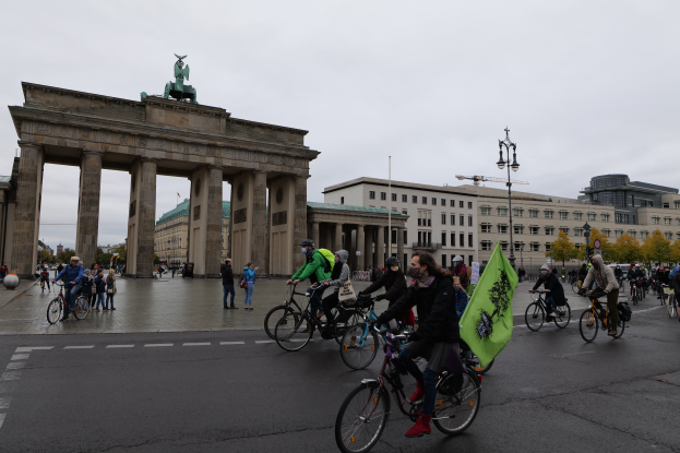 Eine Gruppe von Menschen fährt mit Fahrr√°dern die Stra√če vor dem Brandenburger Tor in Berlin, Deutschland, entlang, einige tragen Taschen, unter einem Himmel mit sichtbaren Laternen und B√§umen.