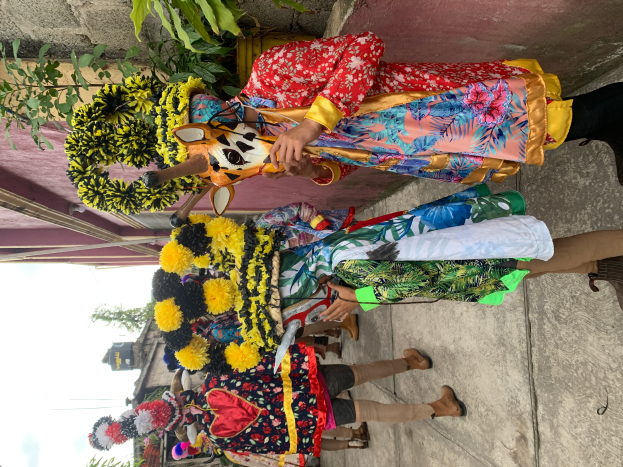 Eine Gruppe von Menschen in bunten Kostümen steht zusammen vor einem Gebäude, mit verschiedenen Gegenständen in den Händen, mit Bäumen, Gebäuden und einem klaren blauen Himmel im Hintergrund während eines Karnevalsumzugs.