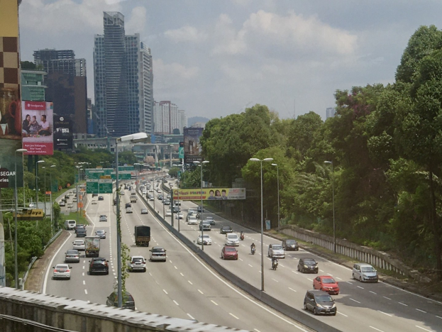 Eine vielbefahrene Autobahn mit hohen Gebäuden, Straßenlaternen, Schildern, Bäumen und Wolken im Hintergrund.