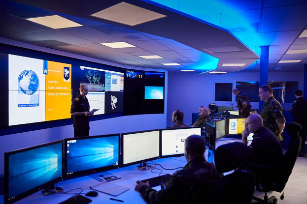 Eine Gruppe von Menschen sitzt an Schreibtischen mit mehreren Monitoren, Tastaturen und Papieren in einer Cyber-Kontrollraum, während eine Person ein Papier hält, mit Bildschirmen und Deckenleuchten im Hintergrund.