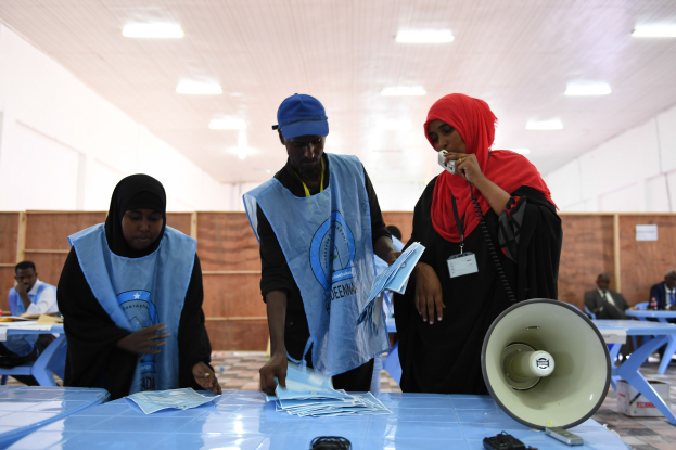 Menschen versammeln sich um einen Tisch mit Megafon und Papieren an einer Wahllokal, mit anderen im Hintergrund sitzend und stehend unter beleuchteter Decke.