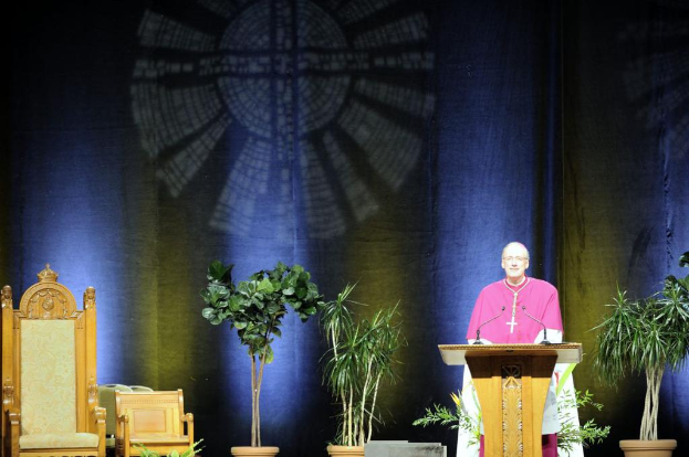 Mann in rosa-weißem Kleid steht neben einem Rednerpult in einem Auditorium mit Pflanzen und zwei Stühlen daneben und einer bunten Wand im Hintergrund.