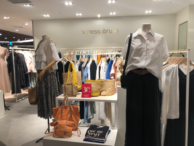 Ein Vanessa Bruno Geschäft in Singapur mit Schaufensterpuppen in Kleidern, hängenden Kleidern, Taschen, einer Texttafel an der Wand, einem Spiegel auf der rechten Seite und Deckenbeleuchtung.