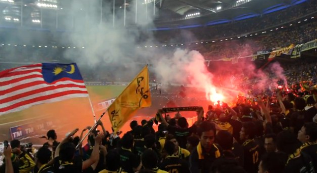 Große Menschenmenge in einem Stadion mit Bengalos, Fahnen, Rauch und Schildern mit der Aufschrift "Malaysia vs Malaysia Live Streaming".