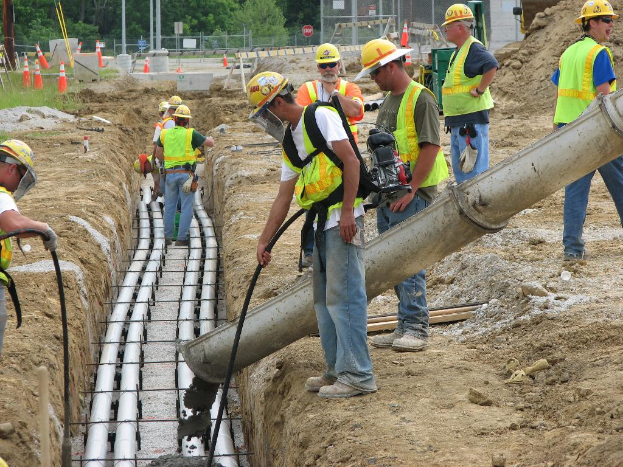 Eine Baustelle mit Arbeitern in Westen und Helmen, Absperrgittern, Zaun, Bäumen, Pfählen, Erde und Rohren im Hintergrund.
