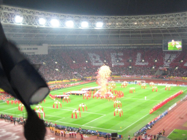 Ein Stadion mit Flutlicht, Eisen Gittern, Zuschauern, stehenden Personen, Werbetafeln, Bildschirmen, Überwachungskameras, Fahnen und Dekorationen.