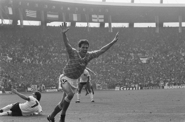 Schwarzes und weißes Foto eines Fußballspielers, der ein Tor feiert, während er von Mannschaftskameraden und Betreuern umgeben ist, mit Tafeln, Flaggen und Pfosten im Hintergrund.