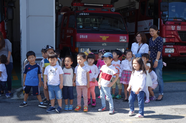 Eine Gruppe von Kindern vor einem Feuerwehrauto auf einem Feuerwehrauto, einige mit Mützen, mit zusätzlichen Feuerwehrautos im Hintergrund.