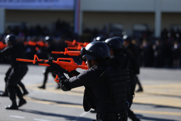 Polizeibeamte in Einsatzausrüstung marschieren auf einer Straße, tragen Helme und halten Waffen, mit einem Gebäude und einer Fahne im Hintergrund.