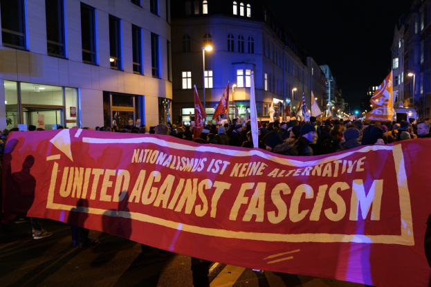 Gruppe von Menschen, die eine Straße entlanggehen und ein Banner mit der Aufschrift 'Einheit gegen Faschismus' tragen, mit Gebäuden, Laternenmasten und Schildern an der Straße unter einem sichtbaren Himmel.