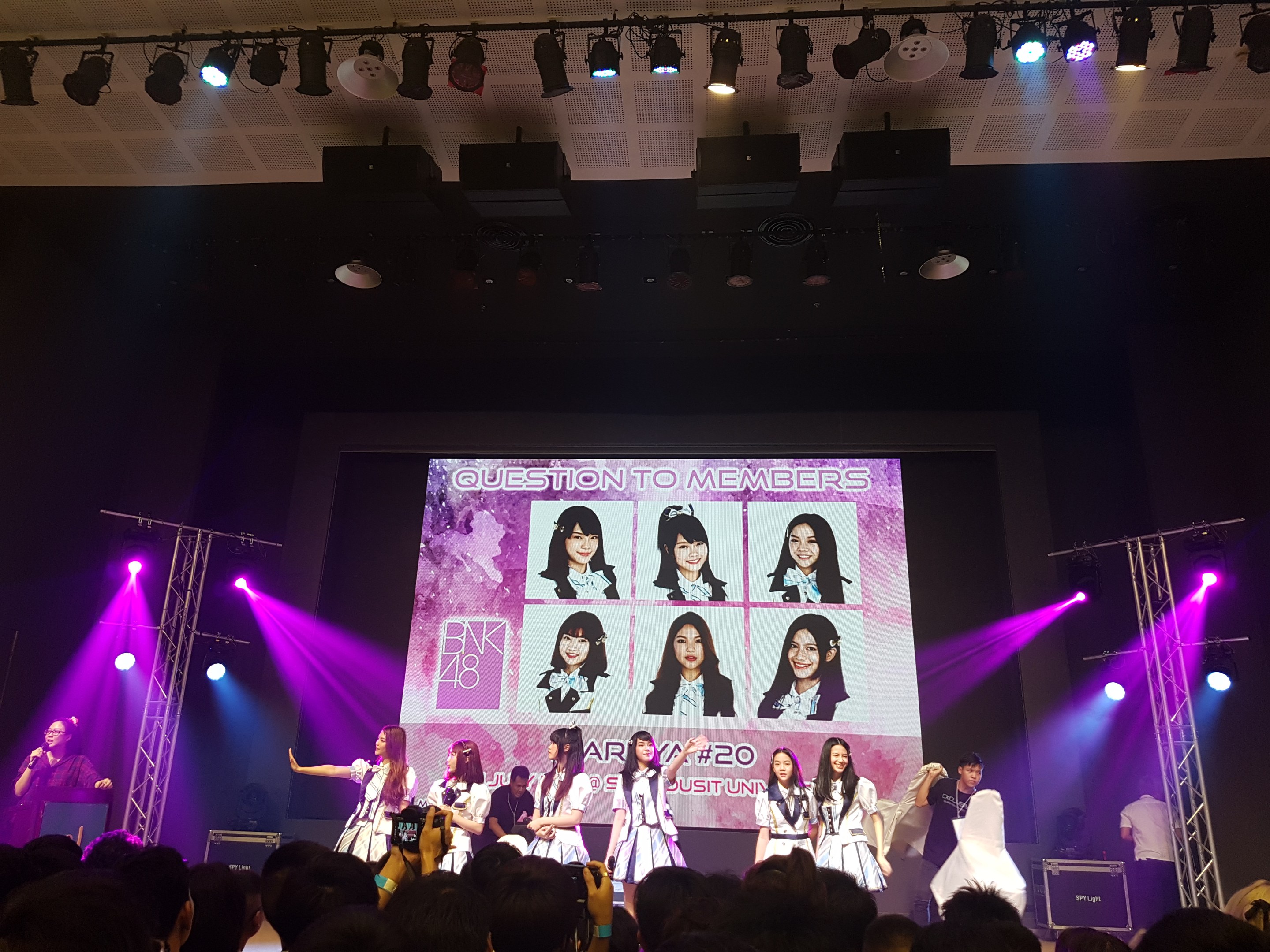 Eine Gruppe von Menschen, die auf einer hell erleuchteten Bühne spielt, mit einer Menge darunter, mit einem Sänger mit Mikrofon und einem Musiker, der ein Instrument spielt, während ein großer Bildschirm Bilder der K-Pop-Gruppe BK48 zeigt.