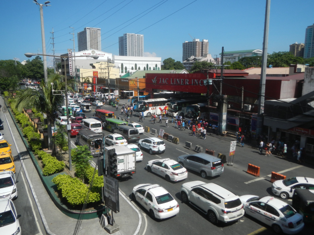 Eine belebte Stadtstraße mit Autos, Bussen, Fußgängern, Bäumen, Laternenmasten, Strommasten, Gebäuden und einem klaren blauen Himmel.