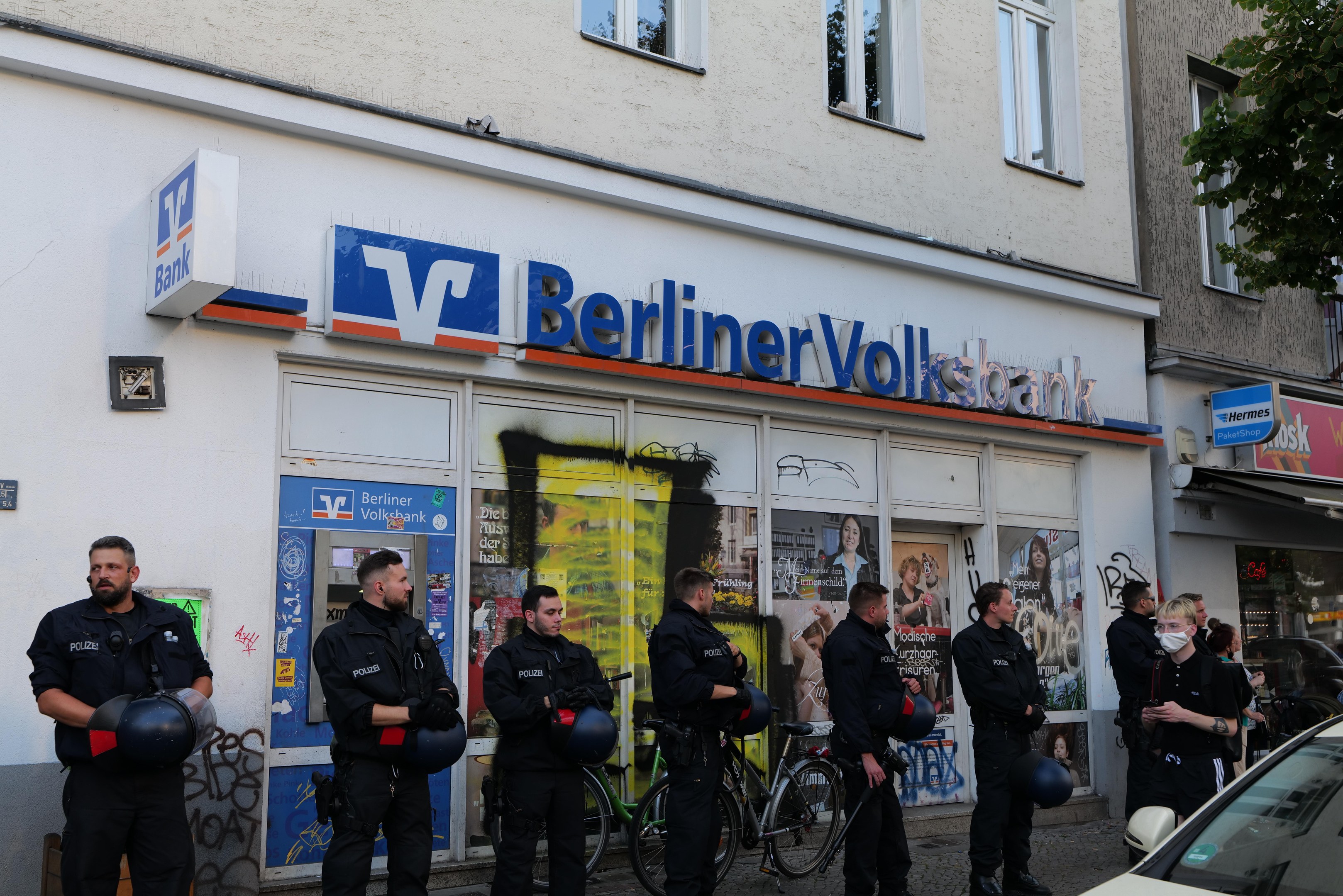 Polizisten vor einer Berliner Volksbank stehend, einige mit Helmen in den Händen, mit einem Auto auf der rechten Seite und einem Baum auf der linken Seite, Gebäude und Texttafeln im Hintergrund.