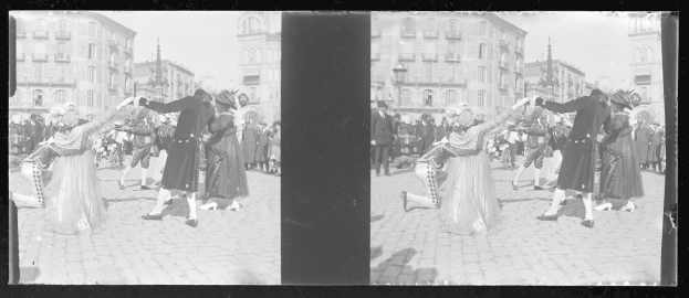Ein Schwarz-Weiß-Foto einer Gruppe von Menschen, die auf der Straße tanzen, während eines Faschingsumzugs, gekleidet in Kostüme, mit Gebäuden, Straßenlaternen und -masten im Hintergrund unter einem klaren Himmel.