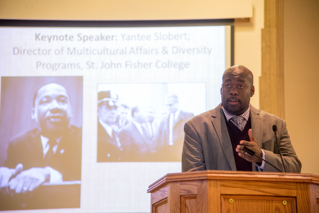 Ein Mann in Mantel, Krawatte und Hemd spricht nahe einem Rednerpult auf der rechten Seite des Bildes, mit einem Projektionsschirm in der Mitte und einem Mann in Schwarz und Weiß auf der linken Seite.
