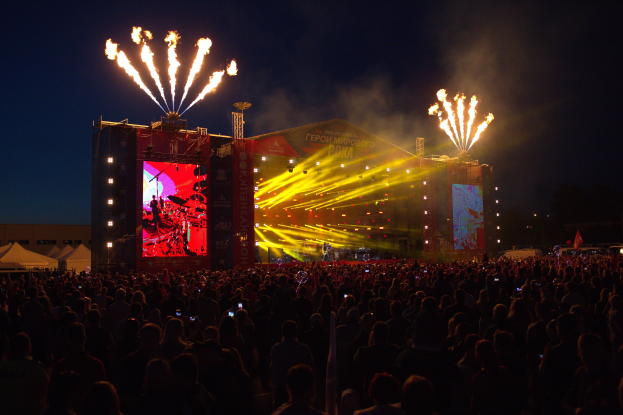 Eine große Menschenmenge auf einem Musikfestival mit einer beleuchteten Bühne, auf der Künstler, Banner und Zelte zu sehen sind, sowie ein Feuerwerk am dunklen Himmel.