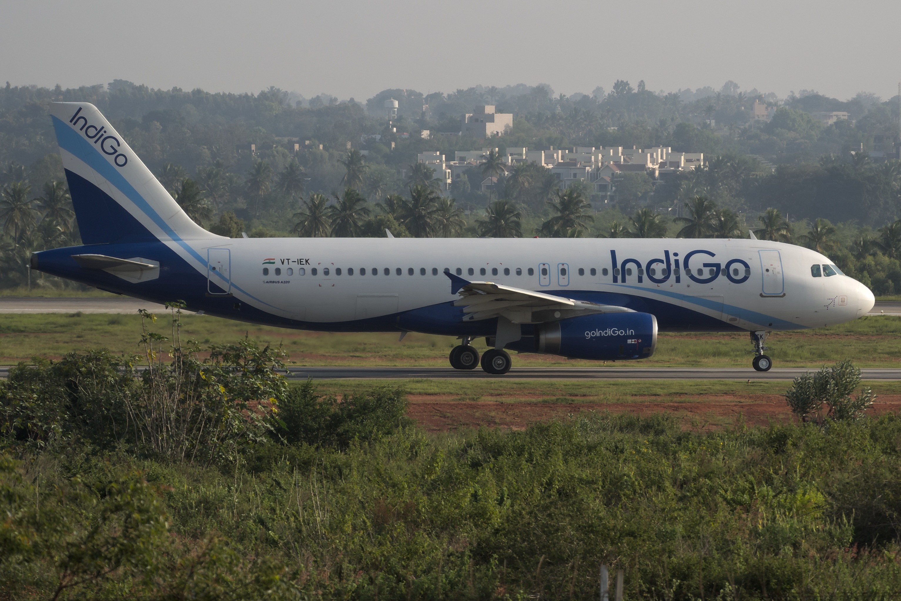 Indigo Airlines Airbus A320-200 auf der Rollbahn am Mumbai Airport mit Grünflächen, Bäumen, Gebäuden und einem klaren blauen Himmel im Hintergrund.