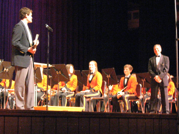 Eine Gruppe von Kindern sitzt auf Stühlen und schaut auf einen Mann in einem schwarzen Anzug, der in der Nähe eines Mikrofons steht, mit einem großen Vorhang im Hintergrund.