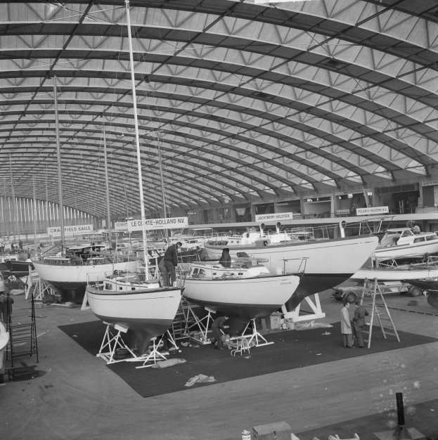 Ein Schwarz-Weiß-Foto von einem belebten Bootshof mit zahlreichen Booten, Menschen bei der Arbeit und verstreutem Equipment wie Leitern und Stäben, mit weiteren Booten im Hintergrund.