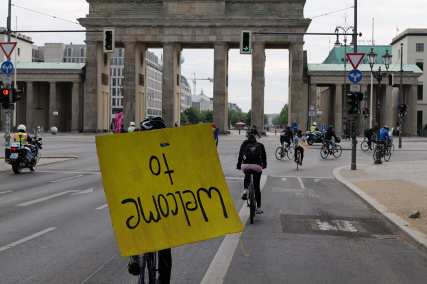 Eine Gruppe von Radfahrern mit Helmen fährt an der Brandenburgertor in Berlin vorbei, einer hält ein gelbes Schild, mit Laternen, Verkehrszeichen, Gebäuden, Bäumen und einem klaren Himmel im Hintergrund.