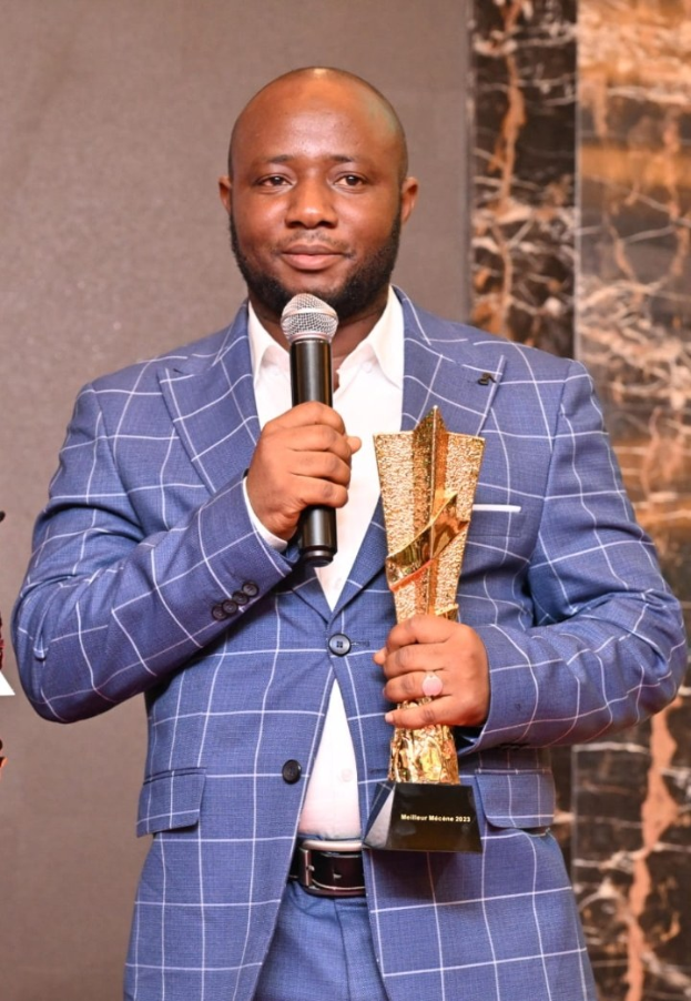 Mann in blauem Anzug mit Mikrofon und Trophäe, feiernd mit einem freudigen Ausdruck und selbstbewusster Haltung.