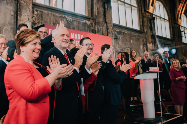 Eine Gruppe von Menschen, die vor einem Publikum klatschen, mit einem Podium, einem Mikrofon und einer Tafel mit Text auf der rechten Seite und Stühlen, einer Fahne, einer Wand, Fenstern und Lichtern im Hintergrund.
