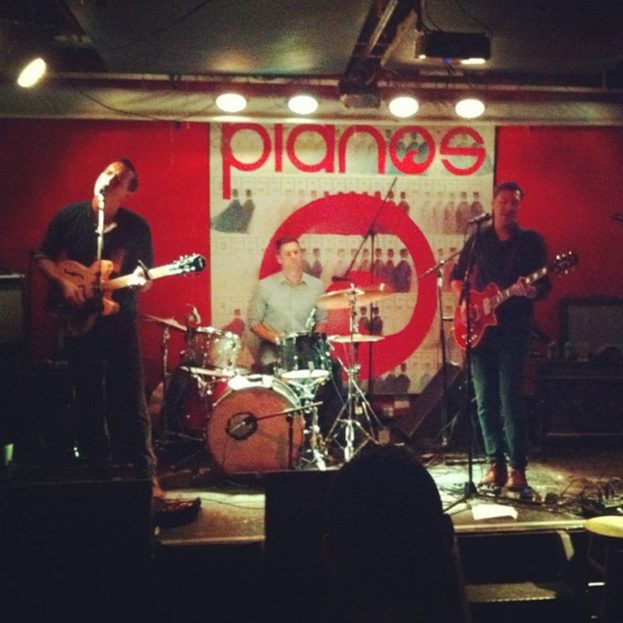 Drei Männer auf der Bühne bei einem Konzert: einer spielt Schlagzeug in der Mitte, und zwei spielen Gitarren und singen auf beiden Seiten, mit einem Banner an der Rückwand und Lampen an der Decke.