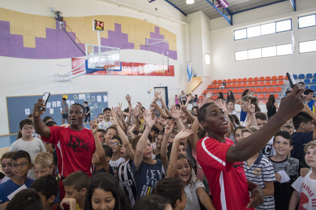 Kinder auf einem Basketballfeld mit Handys stehend, mit einer Anschlagtafel, Uhr, Torpfosten, Basketballkorb, Deckenbeleuchtung, Stühlen und Fenstern im Hintergrund.