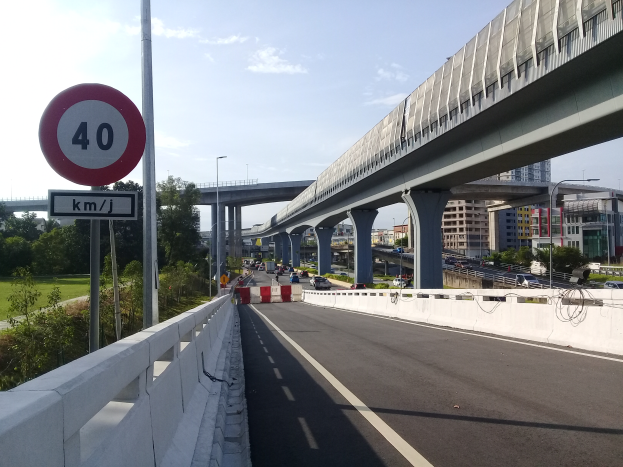 Eine Autobahn mit einem Tempolimit-Schild, Fahrzeugen, einer Brücke mit Pfeilern, Laternen, Bäumen, Gebäuden und einem bewölkten Himmel im Hintergrund.