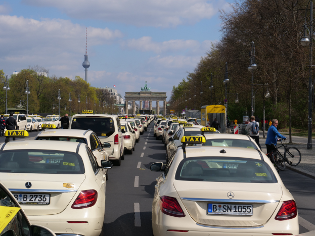Eine lange Reihe von Taxis, die an einer belebten Straße in Berlin, Deutschland, geparkt sind, mit Fahrzeugen, Radfahrern und Fußgängern, flankiert von Laternenmasten, Bäumen und Gebäuden, einschließlich eines Bogens und eines Turms.