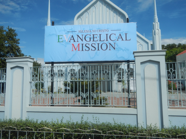 Eine Kirche mit einem Schild, auf dem "Evangelische Mission" steht, ist von einem Metallzaun, Pflanzen, Bäumen und einem Haus mit einem Dach umgeben.