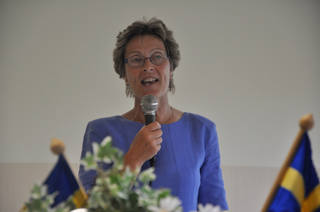 Eine Frau steht an einem Rednerpult mit zwei Flaggen dahinter und spricht in ein Mikrofon.