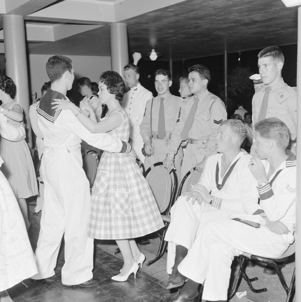 Schwarz-weiß-Bild von Militärpersonal, das auf einer Tanzfläche tanzt und sich vergnügt, mit Säulen und Beleuchtung im Hintergrund.