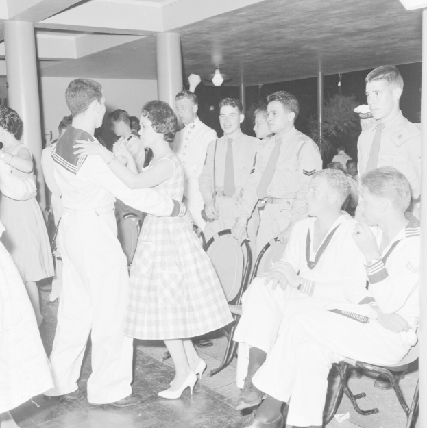 Schwarz-weiß-Bild von Militärpersonal, das auf einer Tanzfläche tanzt und sich vergnügt, mit Säulen und Beleuchtung im Hintergrund.