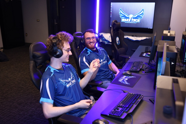 Zwei Männer in blauen T-Shirts und Kopfhörern spielen ein Videospiel an einem Schreibtisch mit Monitoren, Tastaturen und anderem Equipment, während Taschen links auf dem Boden ruhen.