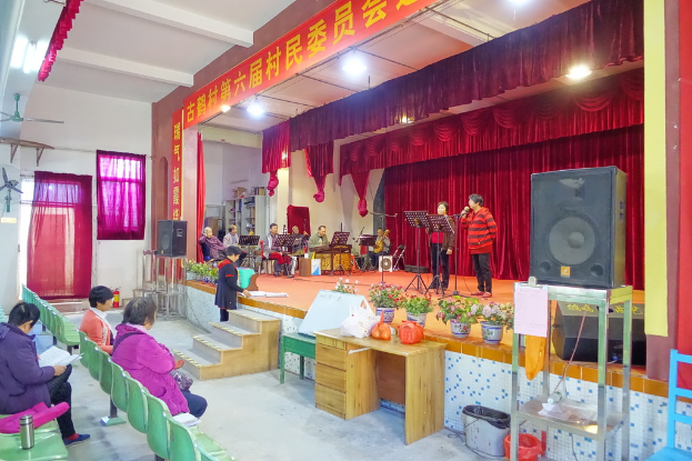 Eine Bühne mit zwei singenden Personen, eine Band im Hintergrund, rote Vorhänge, eine Lampe, ein Lautsprecher auf der rechten Seite und ein Publikum auf Stühlen sitzend.