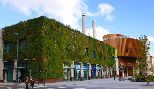 Ein große Bauwerk mit einer grünen Wand an der Seite, umgeben von Straßeninfrastruktur, Fahrzeugen, Füpfördern, Bäumen und einem bewölktem Himmel, als Teil des Green Building Project.