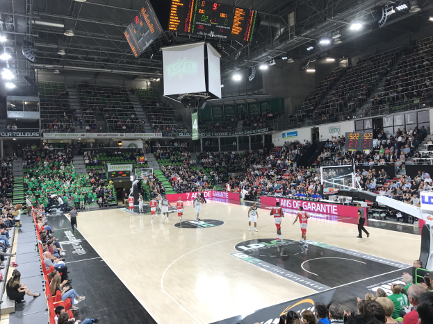 Ein Basketballspiel in einem großen Stadion mit Zuschauern auf den Tribünen und Anzeigetafeln, die 'Asvel vs Galatasaray' oben auf dem Bild anzeigen, unter einer Decke mit Lampen und einem Bildschirm.