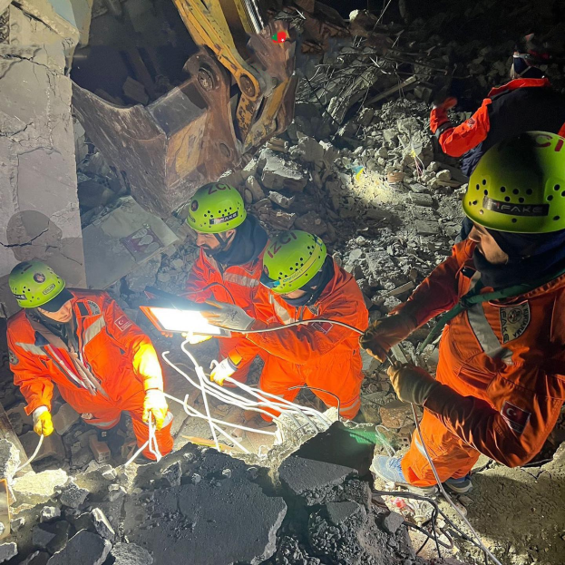 Eine Gruppe von Rettungskräften in orangefarbenen Uniformen und grünen Helmen beseitigt Trümmer von einem eingestürzten Gebäude, unter Verwendung von Werkzeugen, mit einem Kran im Hintergrund.
