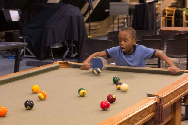 Ein Junge hält einen Tischtennis-Schläger vor einem Snookertisch mit Billardkugeln, mit Stühlen und anderen Gegenständen im Hintergrund.