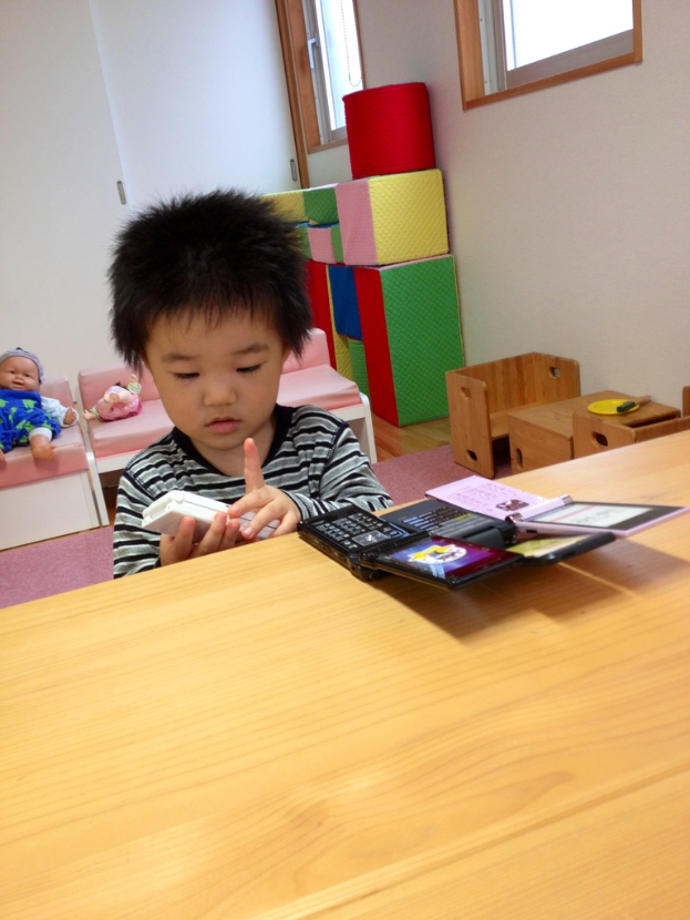 Ein Kind hält ein Mobiltelefon in der Hand mit zusätzlichen Mobiltelefonen auf dem Tisch, eine Puppe dahinter, Stühle, Kisten und Fenster im Hintergrund.