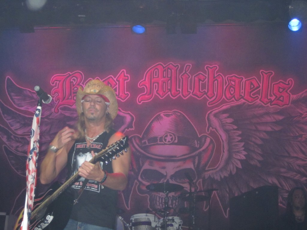 Ein Musikkonzert-Szenario mit Scheinwerfern oben, einem Banner im Hintergrund, Drogen in der Mitte unten, einer Person in der rechten unteren Ecke sitzend und einem Gitarristen mit Mikrofon links.