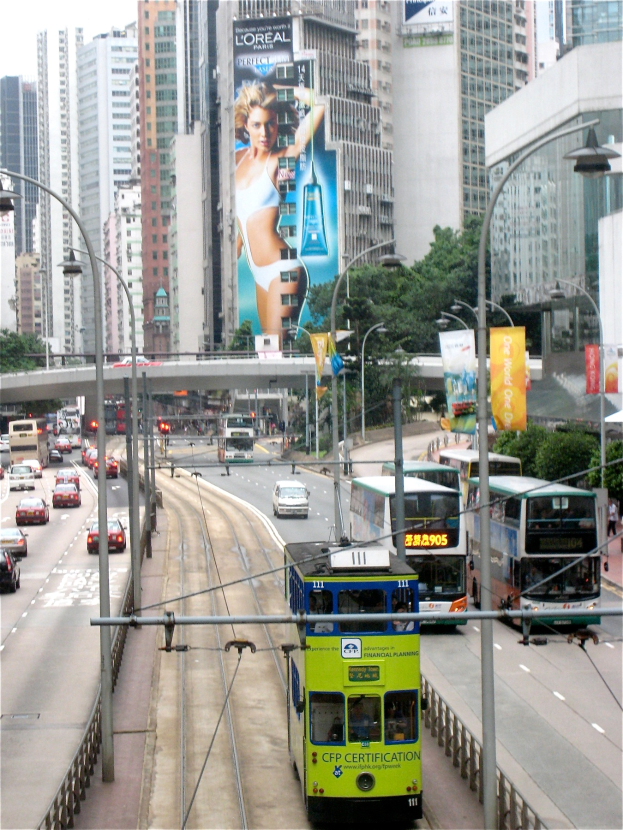Eine belebte Stadtstraße mit verschiedenen Fahrzeugen, einschließlich Doppeldeckerbussen, Autos und anderen Fahrzeugen, umgeben von hohen Gebäuden, Polen, Laternen, Schildern, Bannern, Bäumen und einer Brücke unter einem klaren blauen Himmel, mit Text am unteren Rand des Bildes.