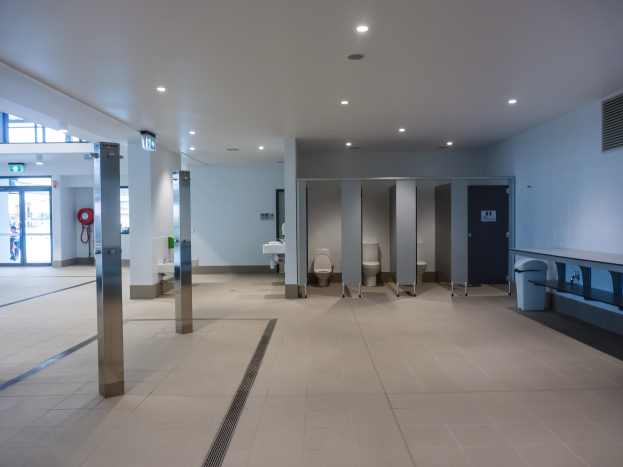 Große öffentliche Toilette mit mehreren Toiletten, tragenden Säulen, einem Müllbehälter, einer Tür, einem Schild, einem an der Wand montierten Feuerlöscher und Deckenleuchten auf dem Dach.