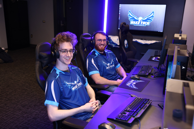 Zwei Männer mit Headsets sitzen an einem Schreibtisch mit Monitoren, Tastaturen und anderem Equipment und lächeln, während sie ein Spiel in einer scheinbar Gaming-Setup mit einem Bildschirm und Lichtern im Hintergrund spielen.